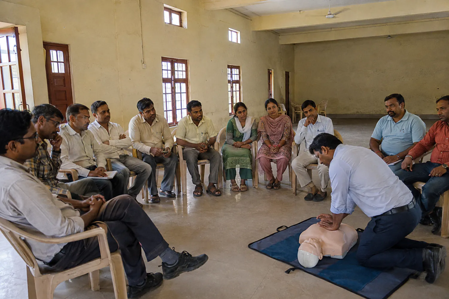 Illustration: a first-aid training session with participants seated around an instructor demonstrating CPR